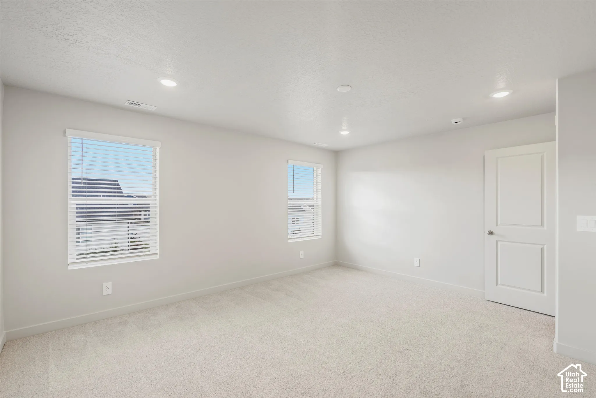 Neutral-toned interior featuring wall-to-wall carpeting, recessed ceiling lighting, and two windows equipped with horizontal blinds