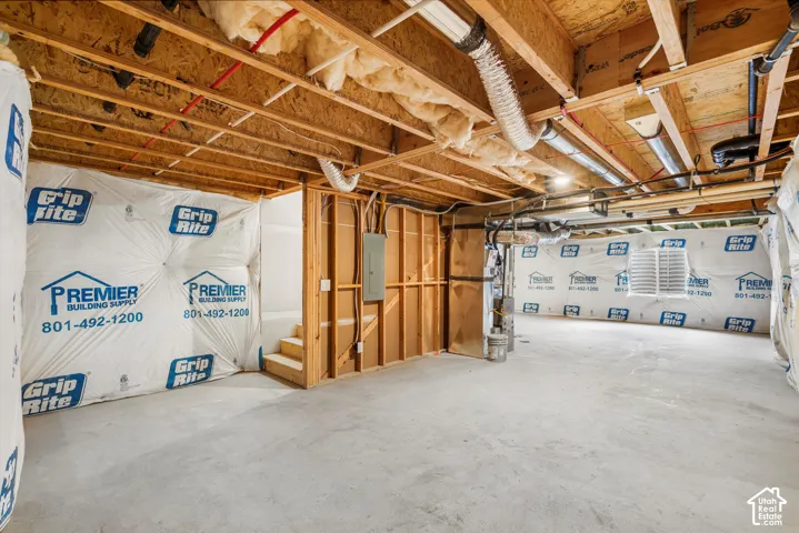 Unfinished lower level featuring concrete flooring, exposed wooden joists, wall-mounted electrical panel, and moisture-barrier foundation wrap