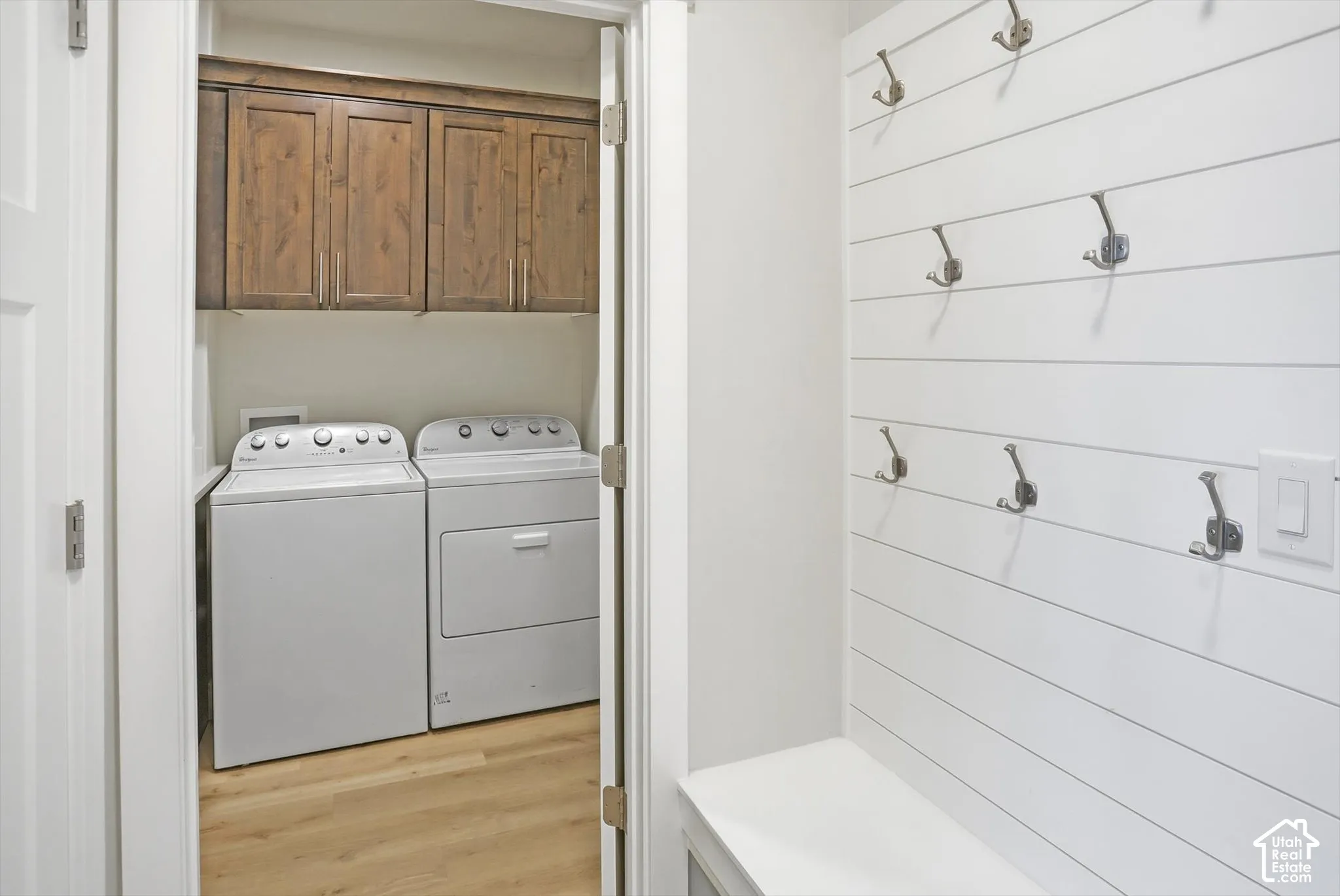 Laundry space featuring wood-finish upper cabinetry and wood-finish flooring, accompanied by a shiplap wall treatment with mounted metal hooks and a built-in bench
