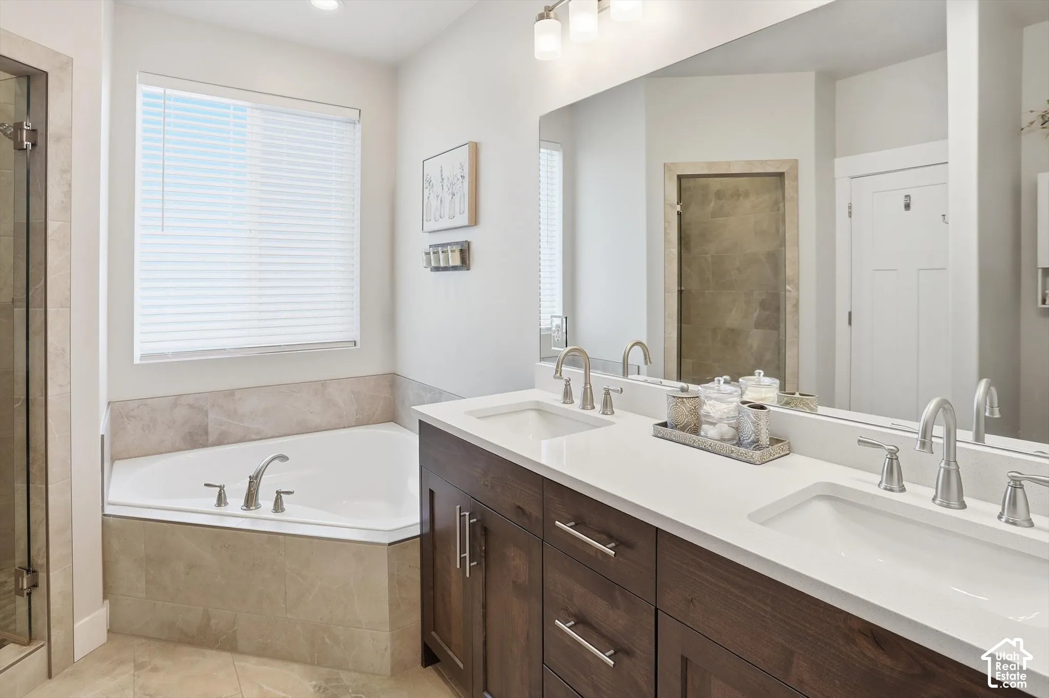 Dual-sink vanity with dark wood-finish cabinetry, solid-surface countertops, soaking tub with stone tile surround, and expansive wall-mounted mirror