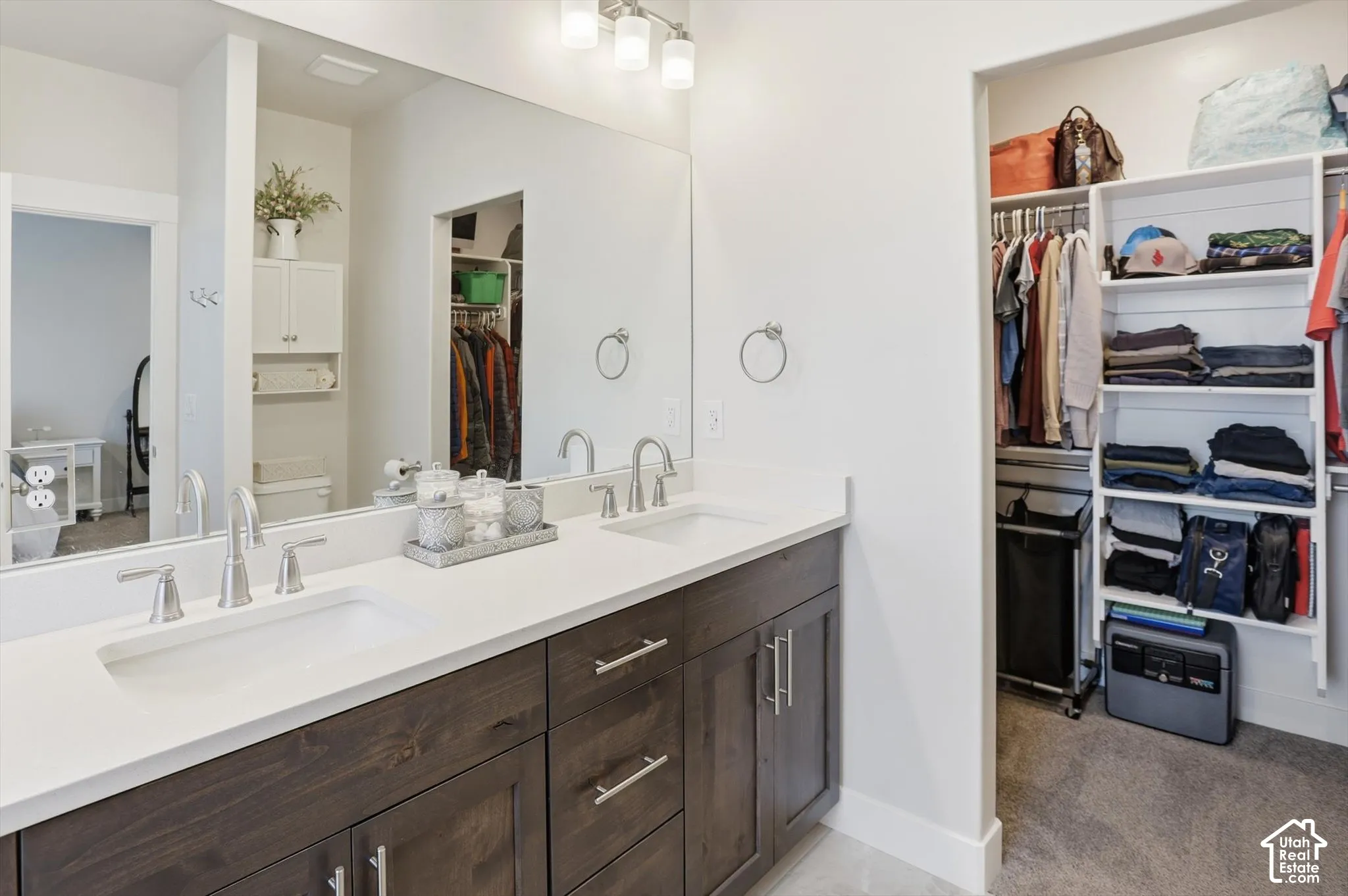 Double vanity featuring a white solid-surface countertop with integrated sinks, dark wood-finish cabinetry, a full-width wall mirror, and an attached walk-in closet with built-in shelving