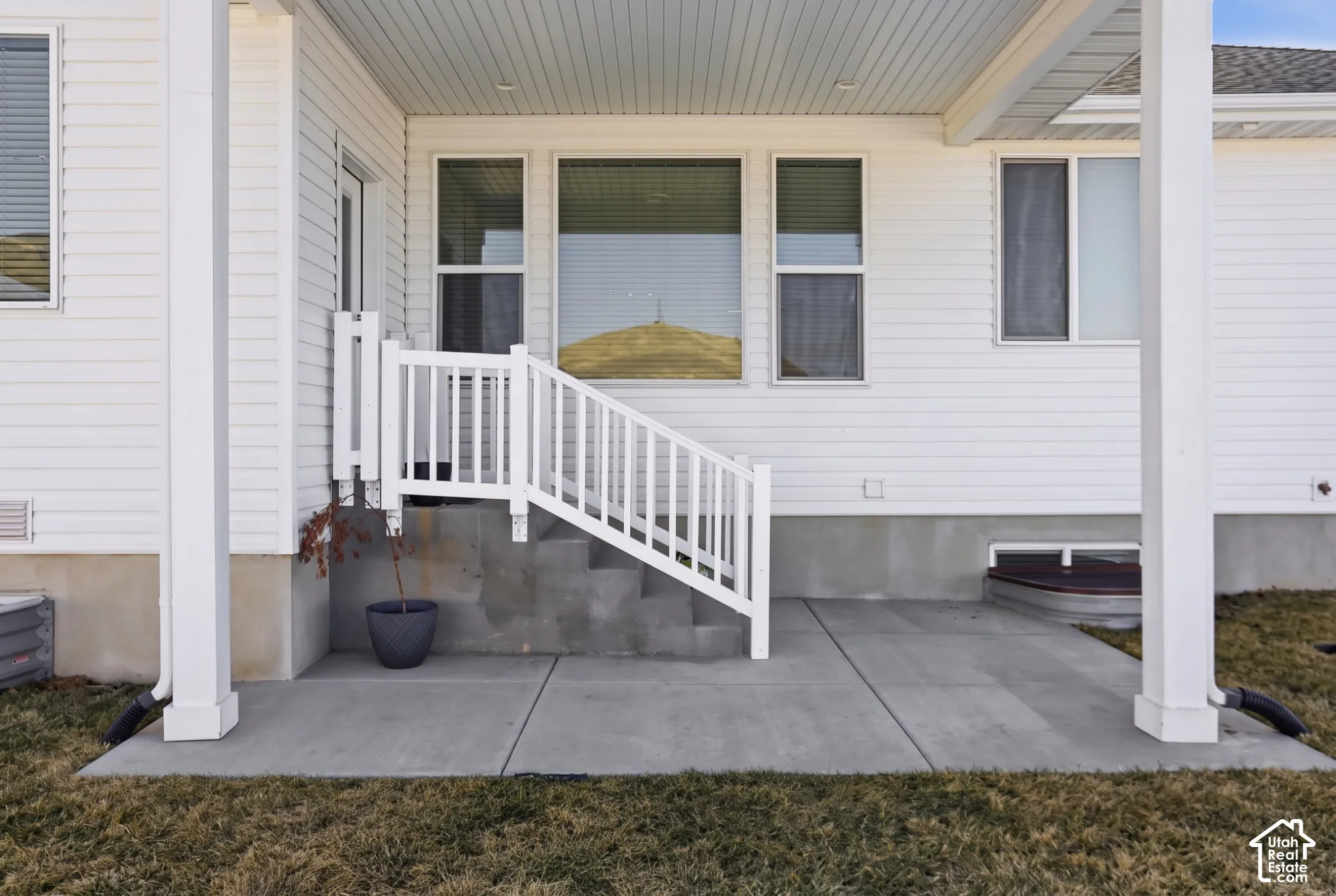 Covered concrete patio featuring white siding, a concrete staircase with white railings, and an egress window well