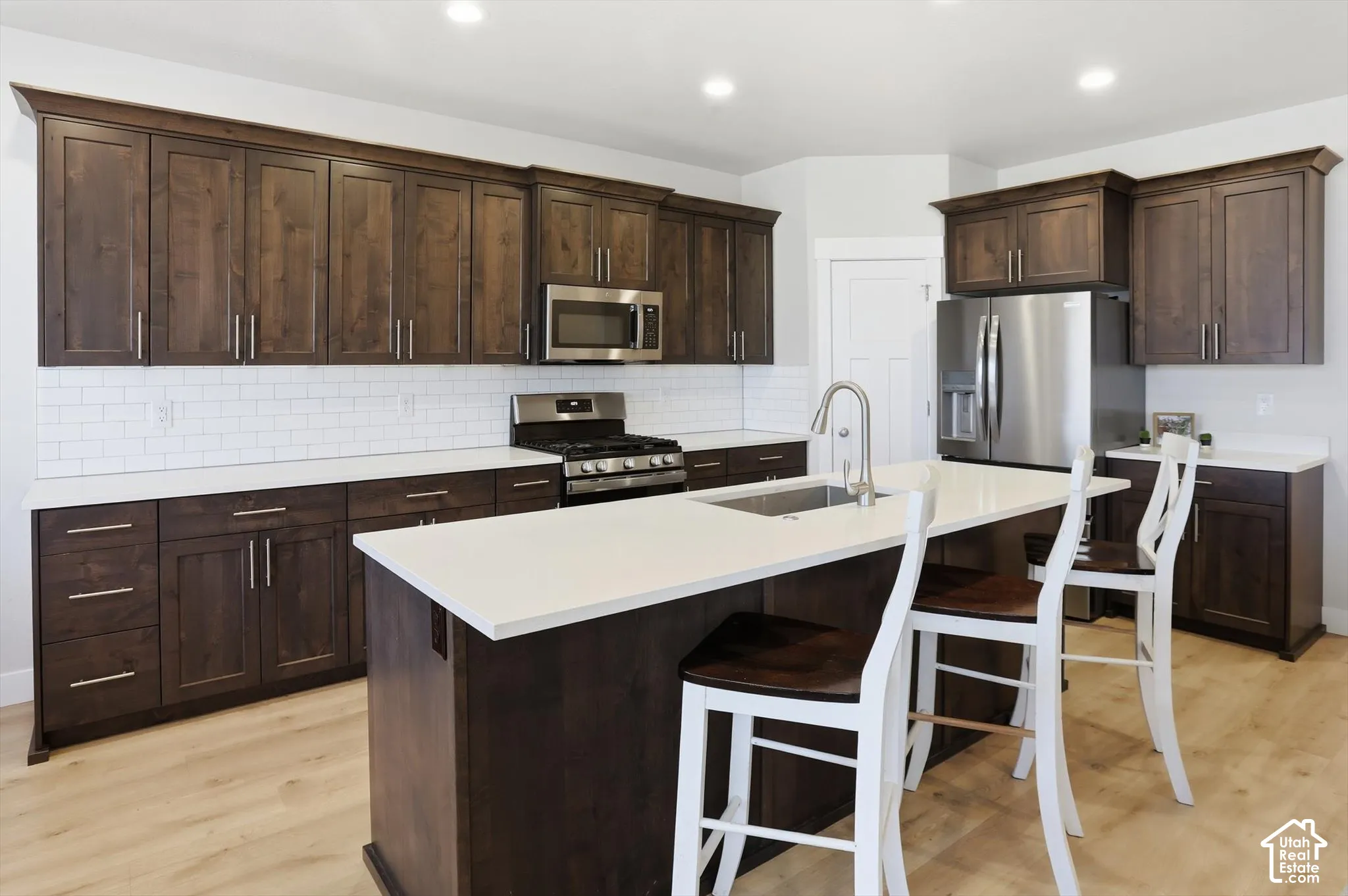 Kitchen featuring dark-finish cabinetry, solid-surface countertops, a center island with an undermount stainless steel sink, a white subway tile backsplash, and stainless steel appliances