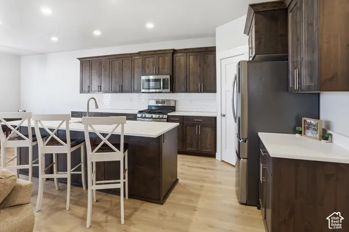 Kitchen featuring dark wood cabinetry, white countertops, a center island with a sink, stainless steel appliances, and a white subway tile backsplash