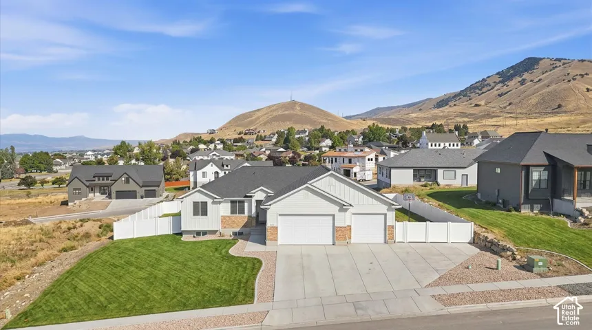 Single-level residence featuring a multi-material exterior of light-toned siding and stone masonry, a three-car garage, white vinyl fencing, and a manicured lawn set against a mountain backdrop