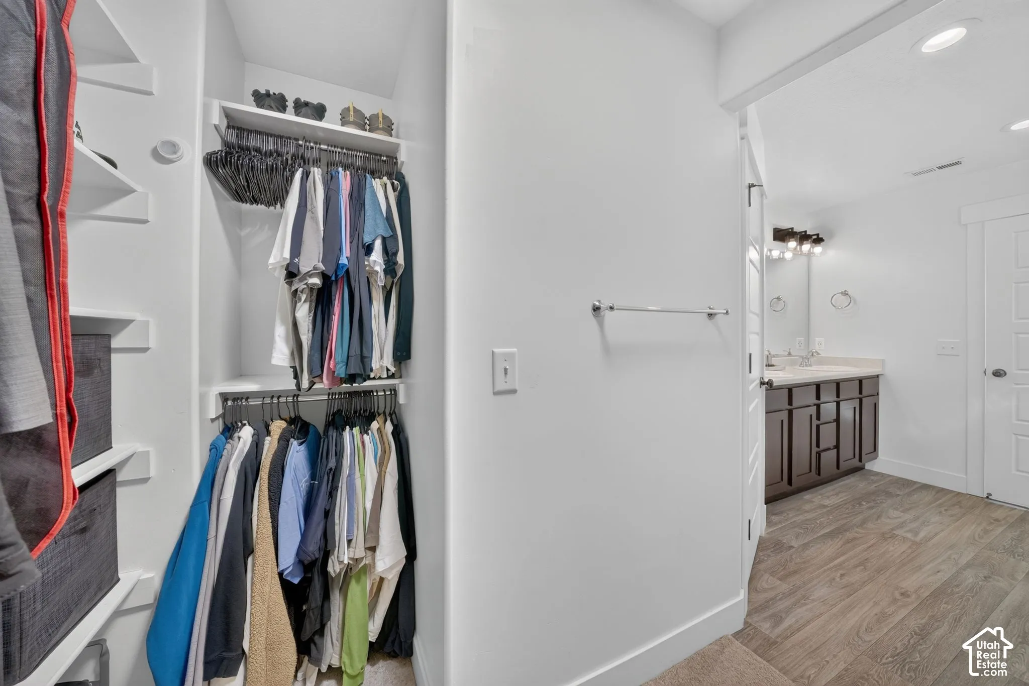 Walk-in closet featuring custom built-in white shelving and dual-tier hanging rods, paired with a vanity area showcasing dark-finish cabinetry, a solid surface countertop, and recessed lighting