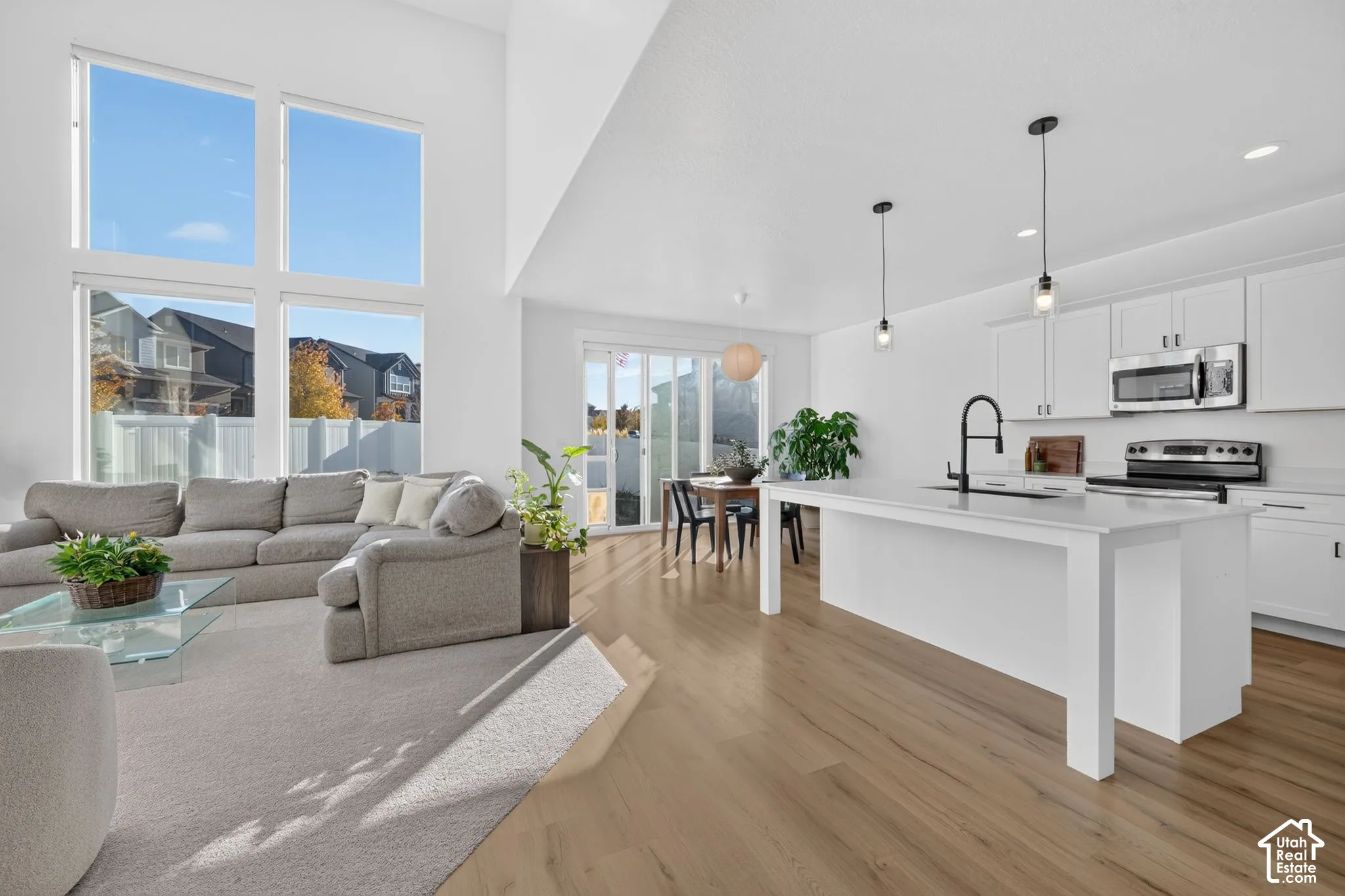 Open-concept area featuring vaulted ceilings, floor-to-ceiling windows, wood-finish flooring, a kitchen island with a neutral countertop, and matte black pendant lighting