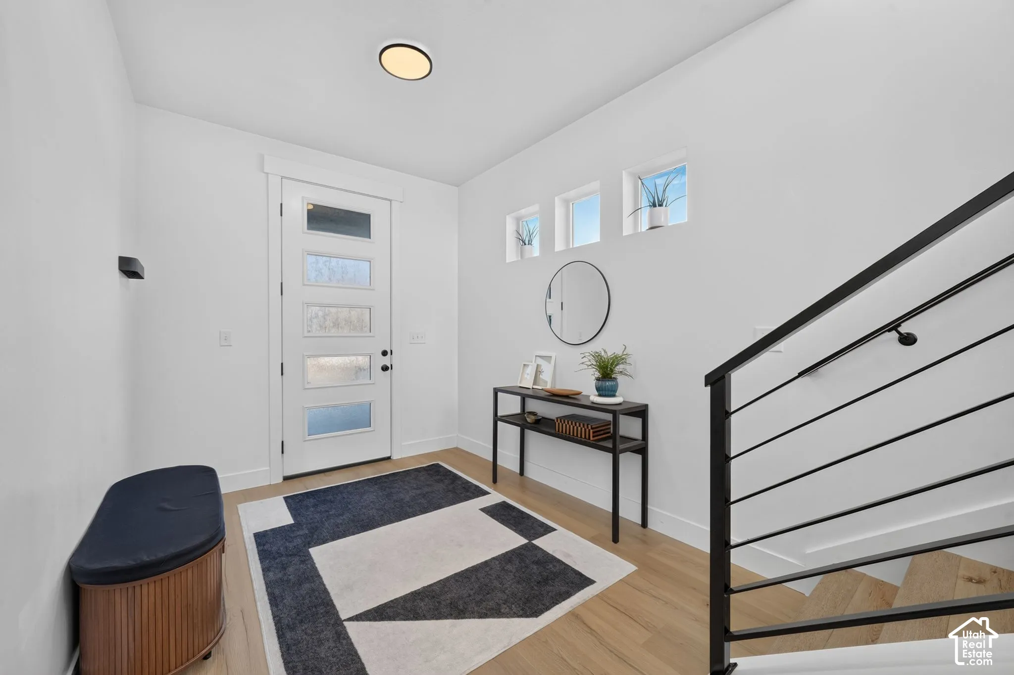 Entryway featuring wood-finish flooring, a contemporary front door with horizontal glass panes, trio of square transom windows, and a black metal staircase railing