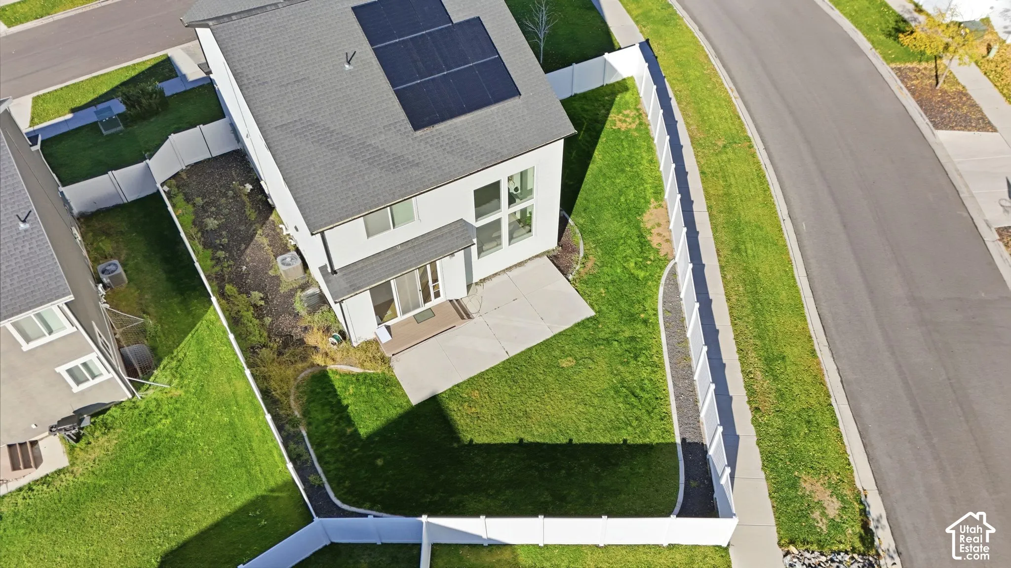 Aerial perspective featuring a multi-story residence with rooftop solar panels, a concrete rear patio, a fenced yard with lawn, and an exterior heating and cooling unit