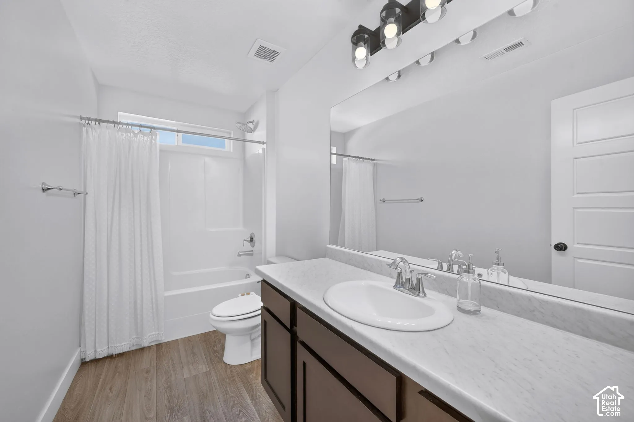Bathroom featuring a wood-finish floor, a vanity with a stone-composite countertop and integrated oval sink, a full-width mirror, a three-light fixture, and a tub-shower combination with an enclosure