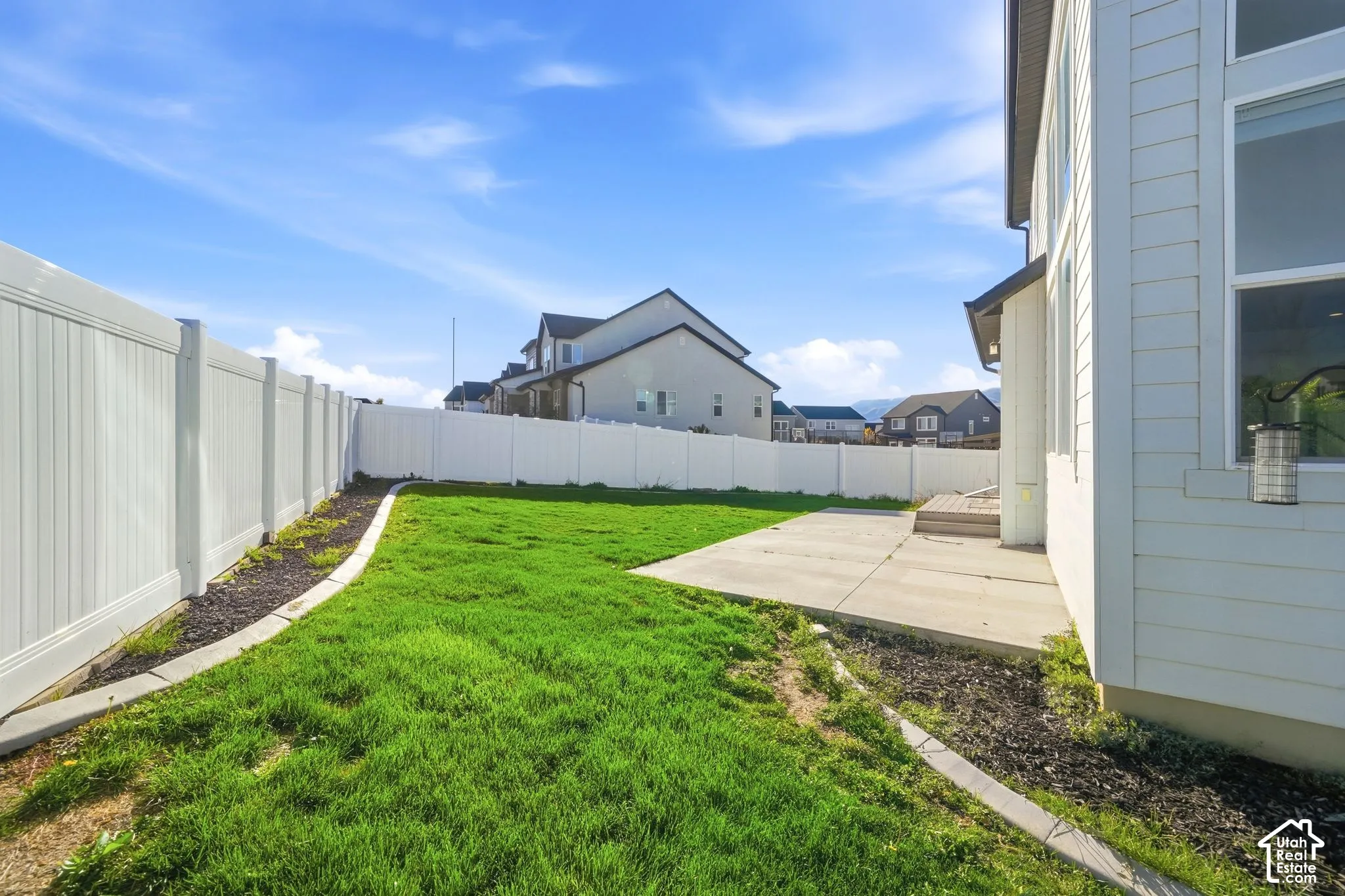 Backyard featuring a concrete patio, white vinyl fencing, and a manicured lawn with landscape curbing and dark mulch beds