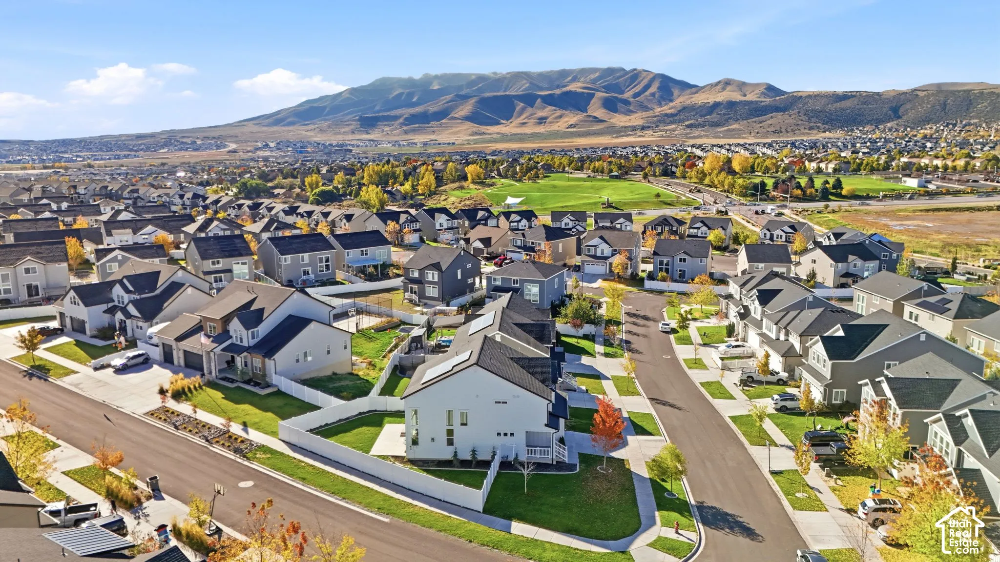 Aerial perspective of a residential neighborhood featuring multi-story homes, paved streets, manicured lawns, white vinyl fencing, and a prominent mountain range backdrop