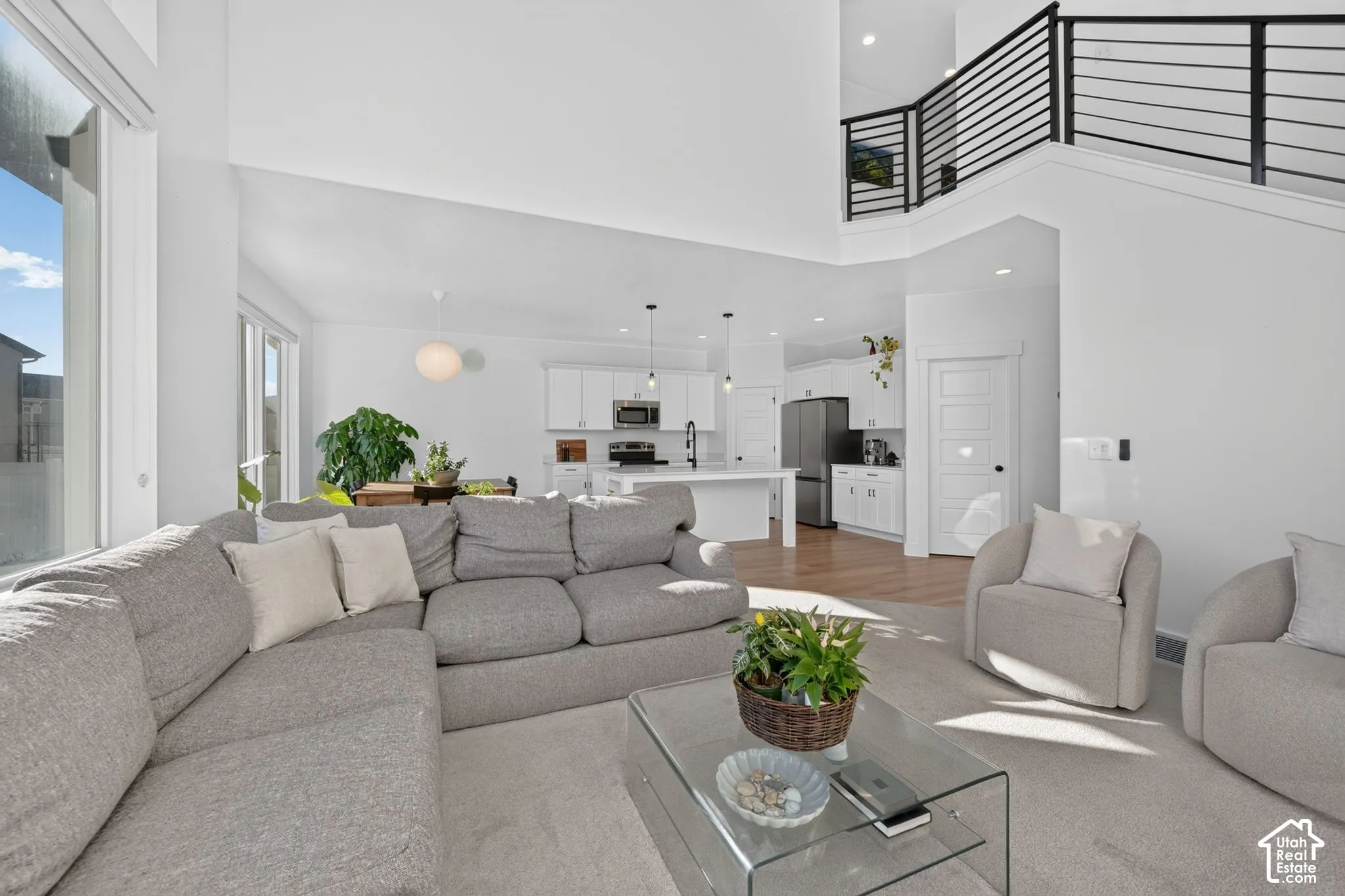 Open-concept interior featuring double-height ceilings, a kitchen with white cabinetry and a central island, recessed lighting, and a metal horizontal-bar staircase railing