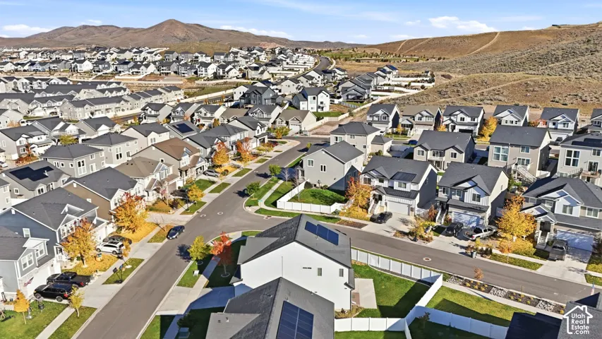 Aerial perspective of a suburban neighborhood featuring residential homes, paved streets, white vinyl fencing, rooftop solar panels, and a backdrop of mountain terrain
