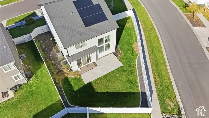 Aerial perspective featuring a multi-story residence with rooftop solar panels, a concrete rear patio, a fenced yard with lawn, and an exterior heating and cooling unit