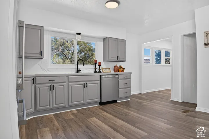 Kitchen featuring gray cabinetry, stone-finish countertops with a full-height backsplash, matte black faucet, integrated stainless steel dishwasher, and wood-finish flooring