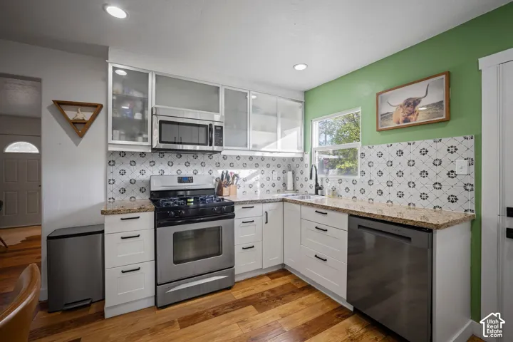Kitchen featuring white shaker-style cabinetry with glass-front uppers, patterned tile backsplash, stone-finish countertops, stainless steel appliances, and wood-finish flooring