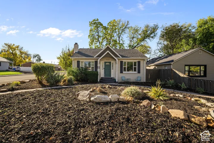 Single-story exterior featuring light-toned masonry, a steep gabled entryway, dark-finish shutters, a decorative stone-lined garden, and an exterior brick chimney