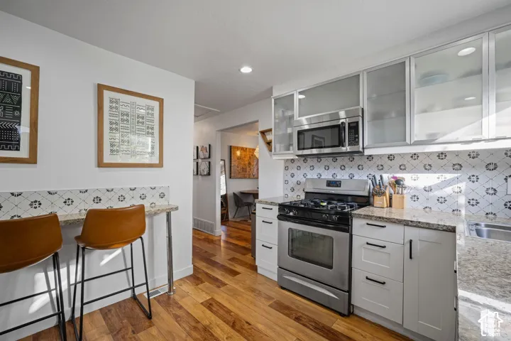Kitchen featuring stone countertops, shaker-style cabinetry with glass-front uppers, stainless steel appliances, patterned tile backsplash, and wood-finish flooring