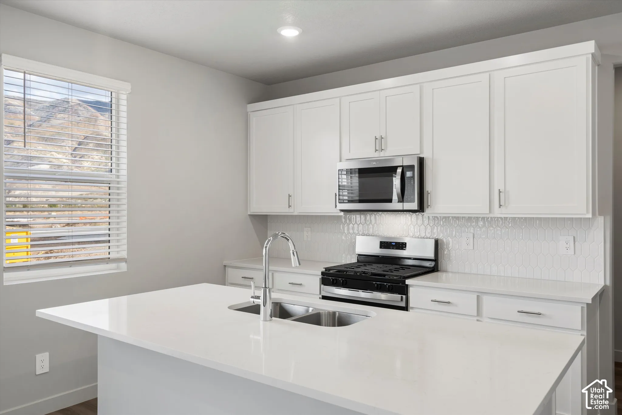 *DISCLAIMER* Interior color package may vary. Kitchen featuring white shaker-style cabinetry, a quartz countertop island with a dual-basin undermount sink, a herringbone-patterned backsplash, and stainless steel appliances