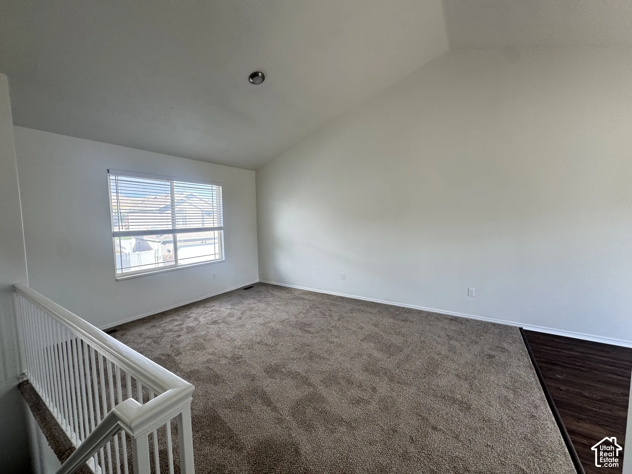 Vaulted ceiling featuring neutral-toned carpeting, a large window with white horizontal blinds, and a white balustrade railing