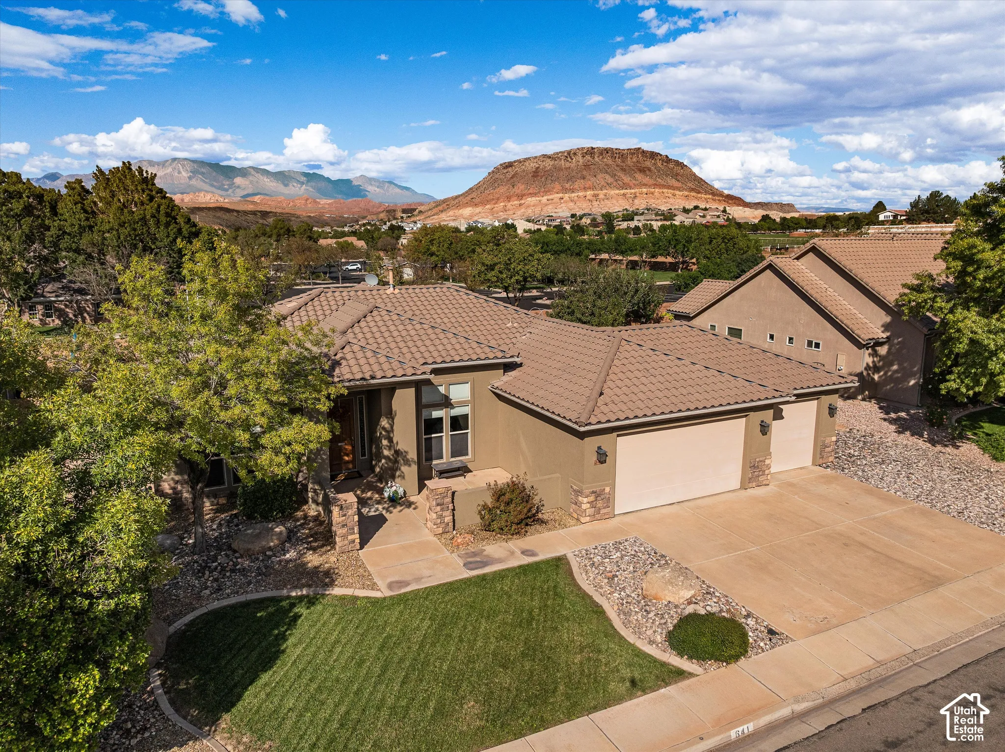 Stucco exterior featuring stone accents, a tile roof, a three-car garage, and a landscaped front yard with a concrete driveway against a mountain backdrop
