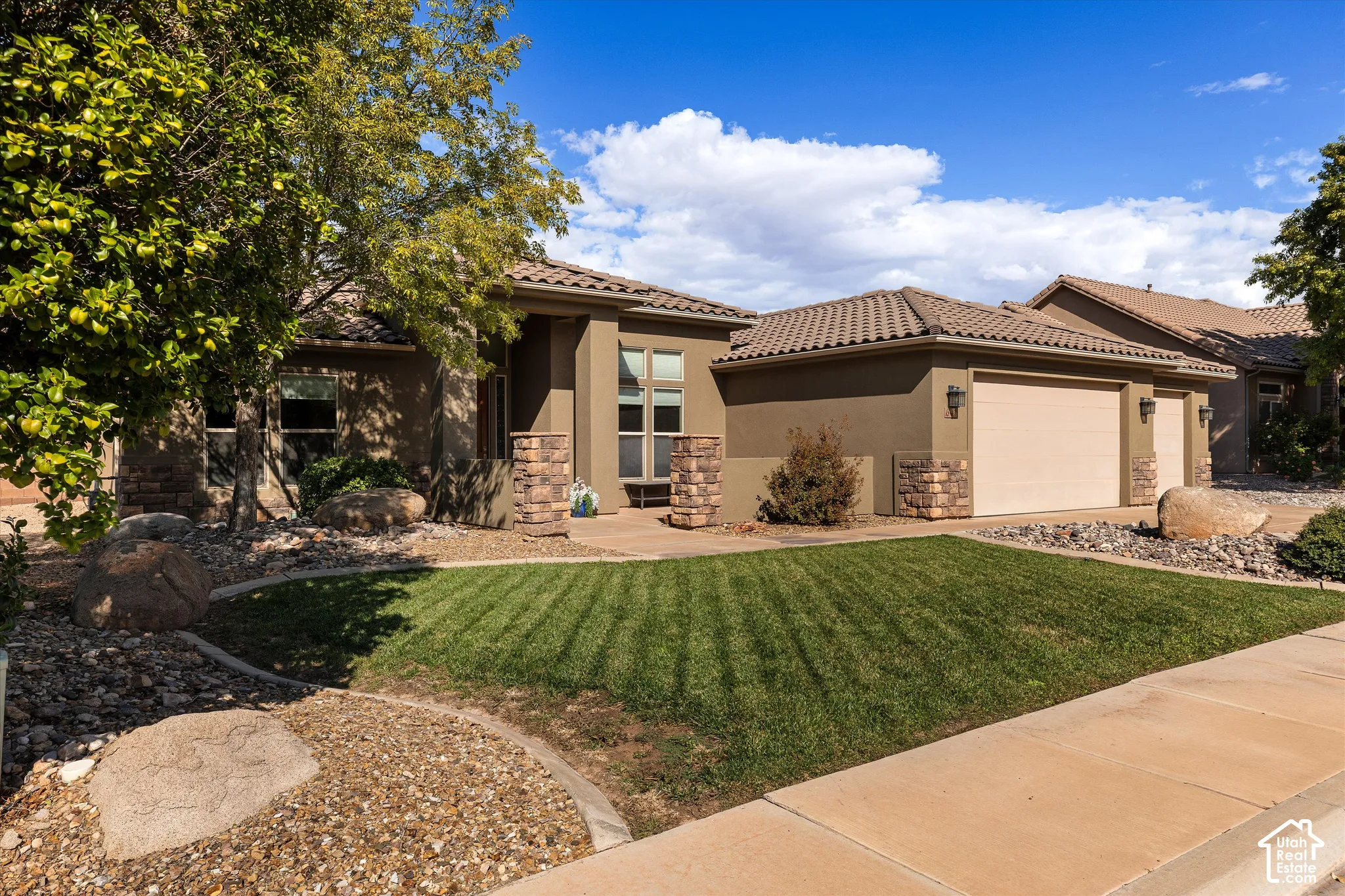 Stucco exterior with stone veneer accents, multi-car garage, concrete tile roof, and landscaped front yard with decorative boulders