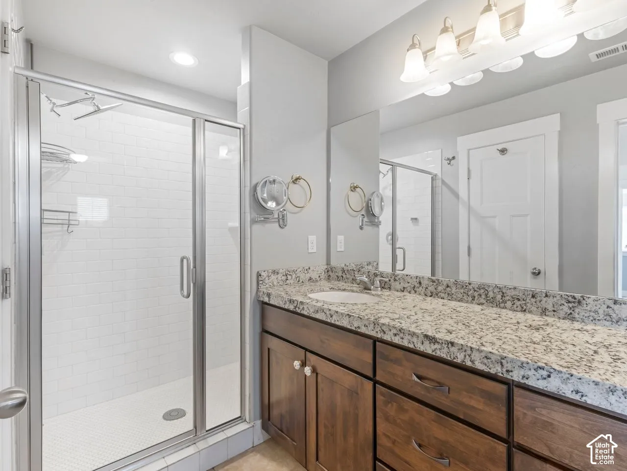 Bathroom featuring a glass-enclosed shower with subway tile surround, stone-finish countertop, dark wood-finish cabinetry, full-width vanity mirror, and multi-bulb overhead lighting
