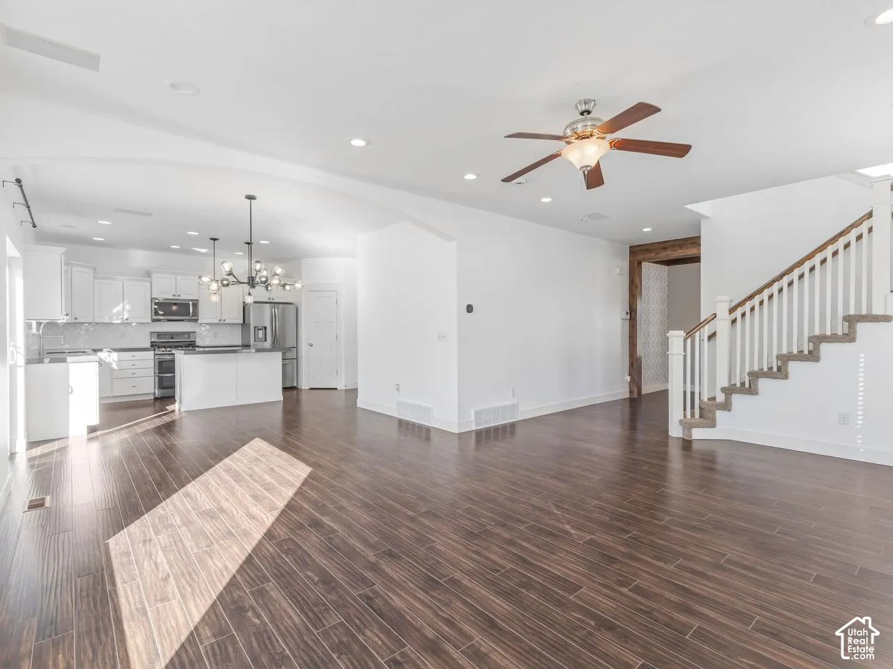 Open-concept interior featuring wood-finish flooring, a kitchen with white cabinetry and an island, a multi-light chandelier, a ceiling fan, and a staircase with a wooden handrail