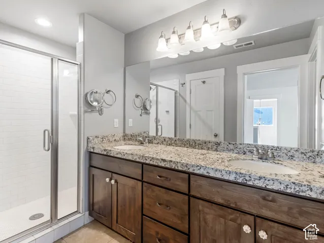 Bathroom featuring a dual-sink vanity with stone countertops, wood-finish cabinetry, a full-width mirror, and a glass-enclosed shower with tiled walls
