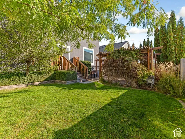 Landscaped backyard featuring a grass lawn, wooden pergola, elevated wooden deck with stairs, and mature trees