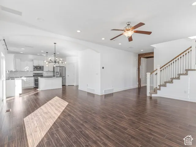 Open-concept interior featuring wood-finish flooring, a kitchen with white cabinetry and an island, a multi-light chandelier, a ceiling fan, and a staircase with a wooden handrail