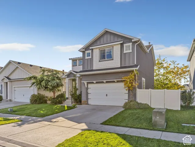 Two-story exterior featuring fiber cement vertical siding, shingle-style accent paneling, a concrete driveway, two-car garage, and white vinyl perimeter fencing