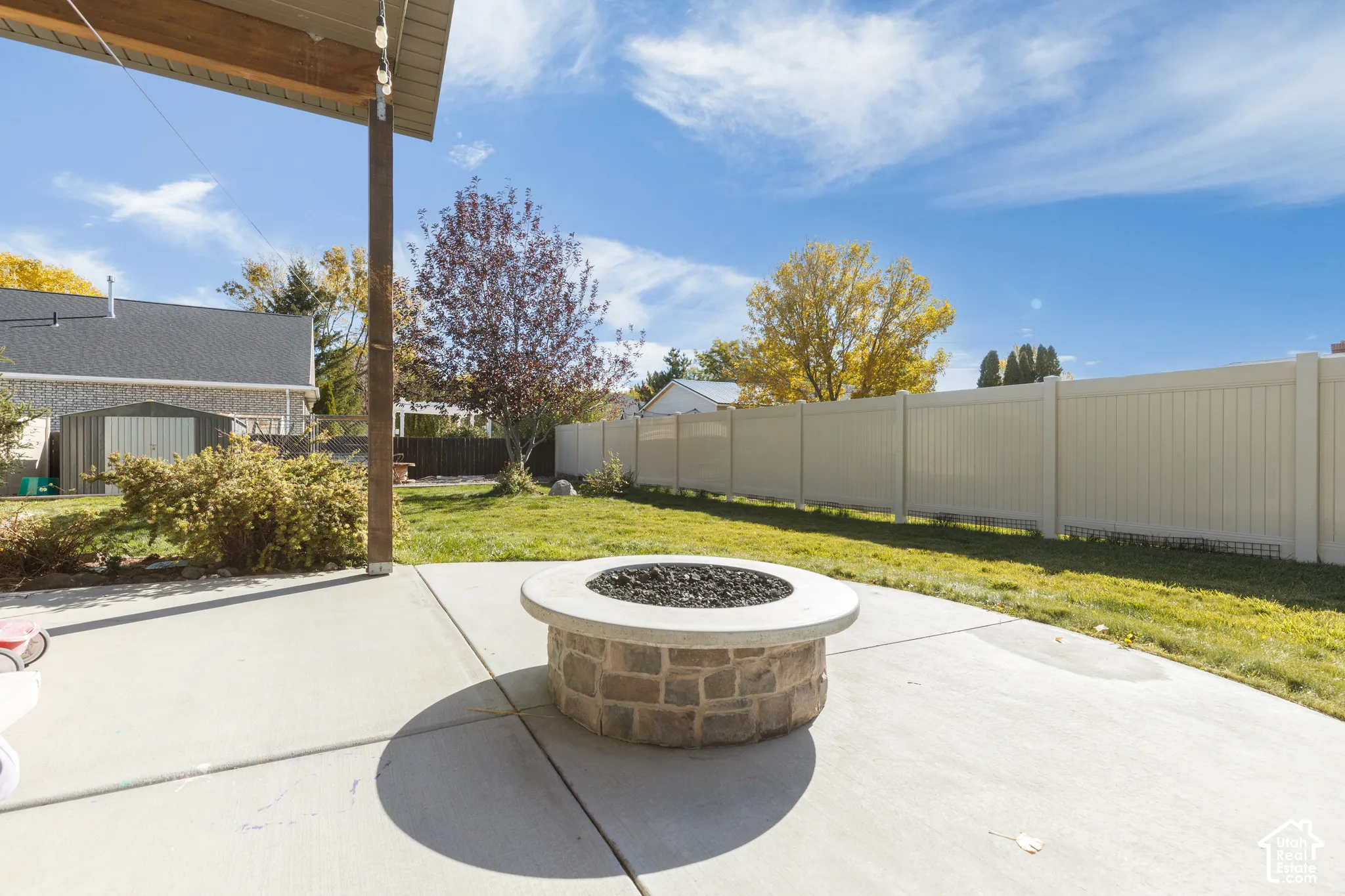 Concrete patio featuring a circular stone-veneer fire pit, covered porch area with wood-finish beam, and fenced backyard with grass lawn