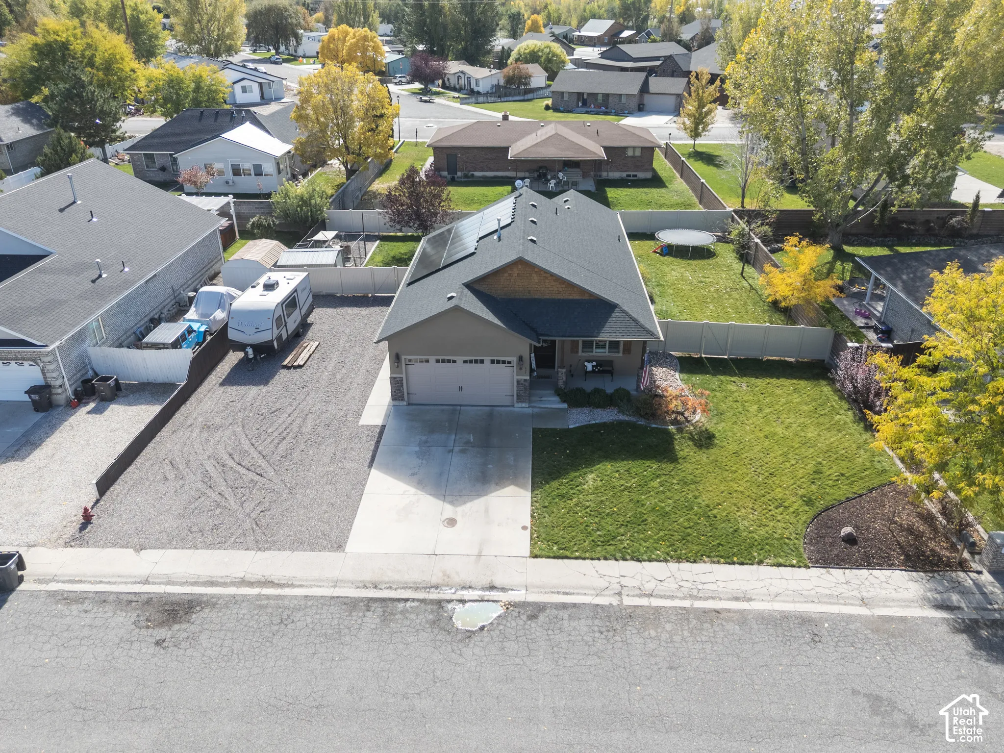 Aerial view of a residential property featuring an attached two-car garage, solar panel array, concrete driveway, gravel side parking area, and fenced lawn