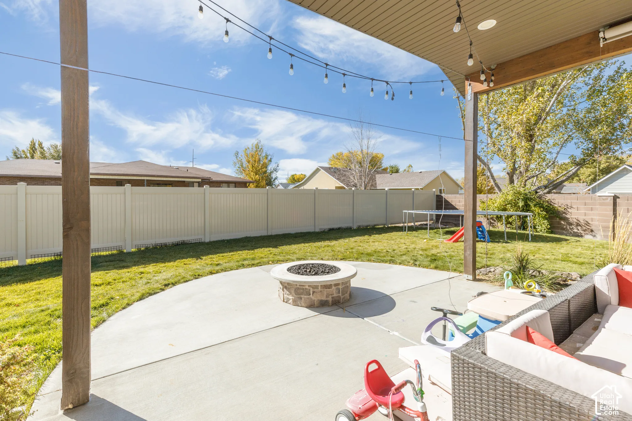 Covered patio featuring a concrete hardscape, stone fire pit, and string lighting, enclosed by a perimeter vinyl fence