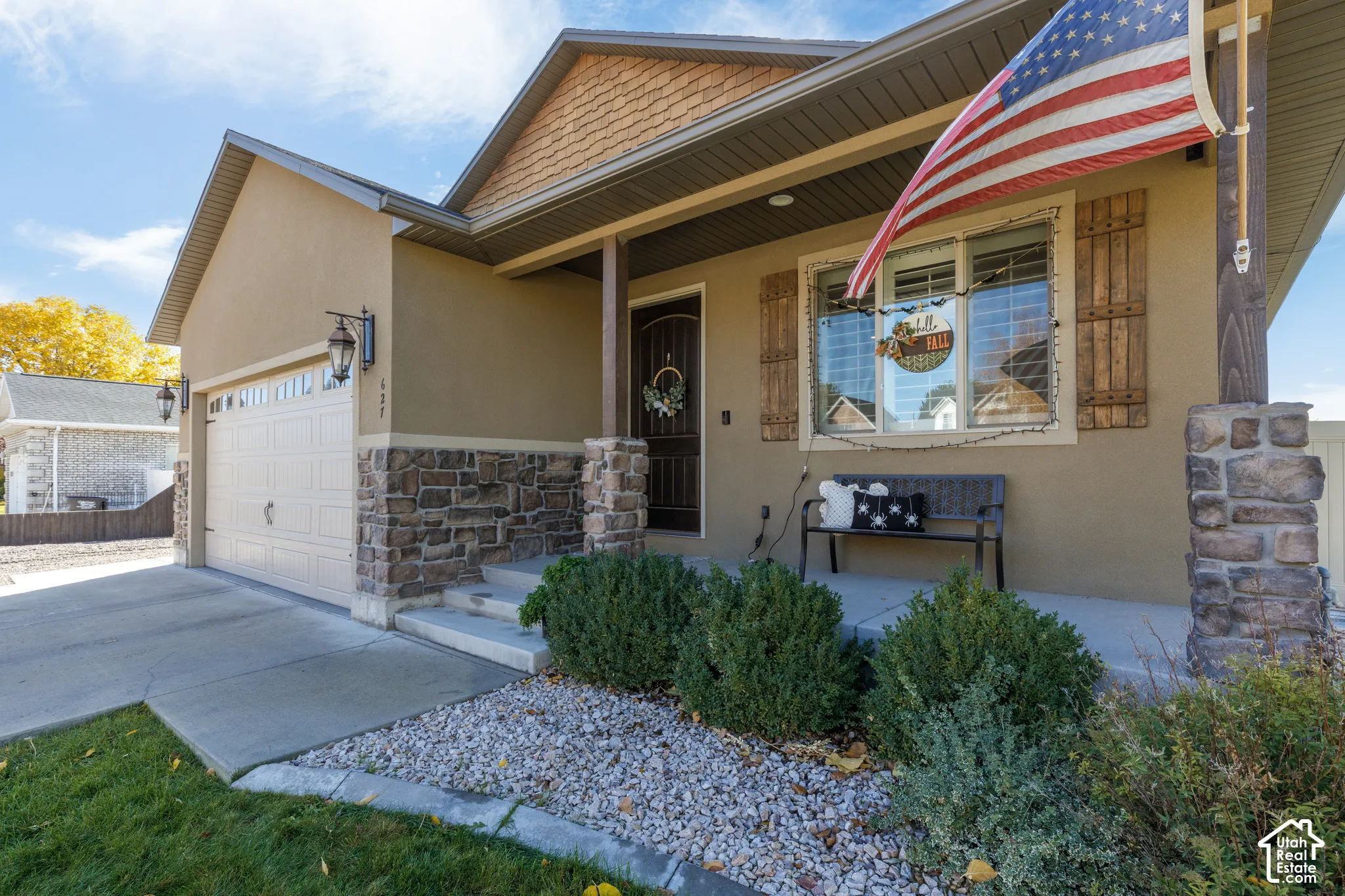 Exterior facade featuring tan stucco siding, stone veneer accents, shingle-style gable detailing, a covered front porch with timber posts, and decorative wooden shutters