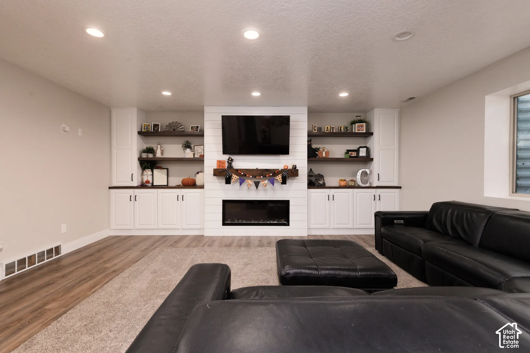 Built-in white cabinetry, horizontal shiplap fireplace surround, recessed lighting, wood-finish flooring, and natural wood floating shelves