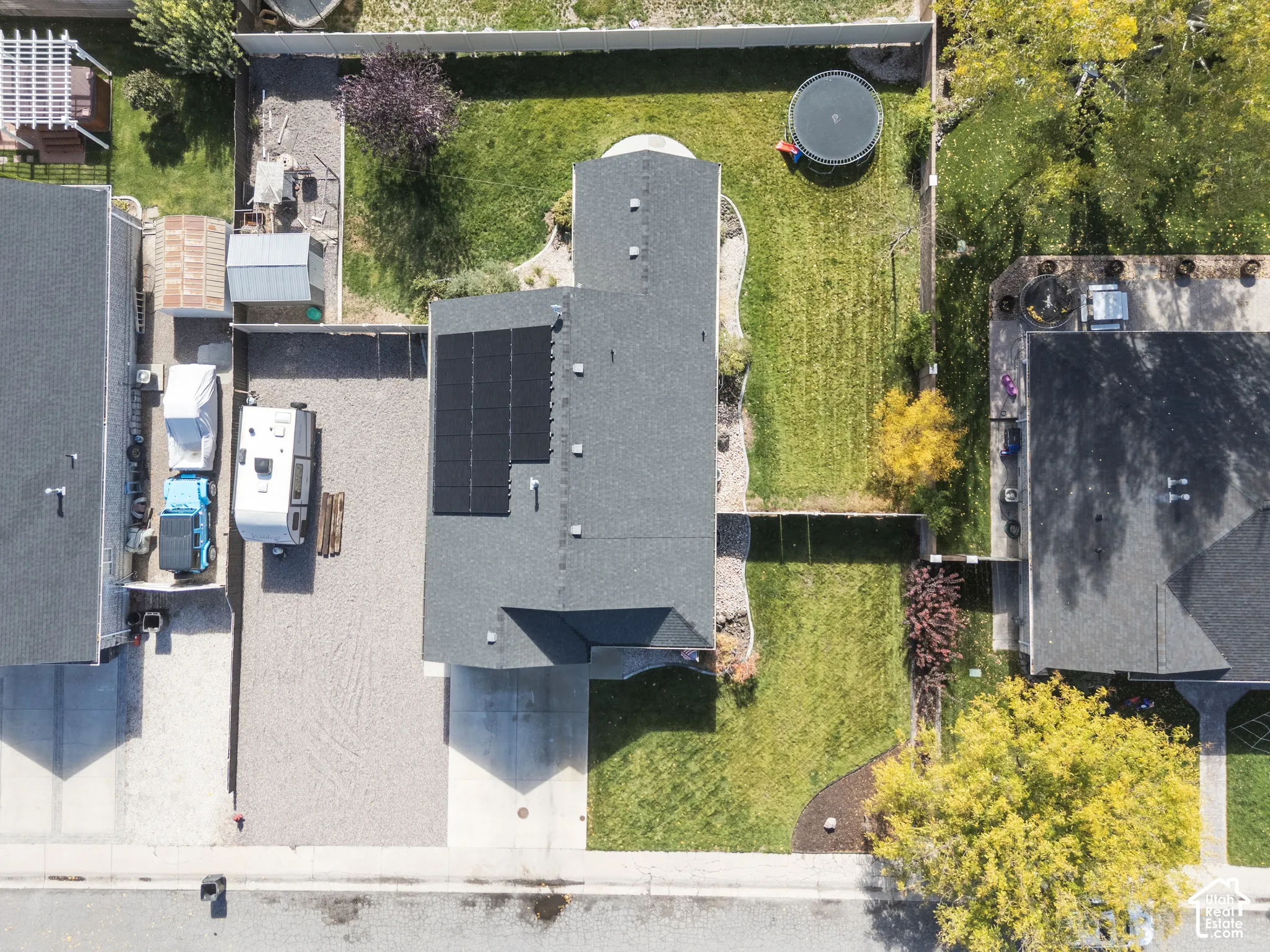 Aerial perspective featuring a shingled roof with integrated solar panels, a fenced backyard, and a gravel side parking area