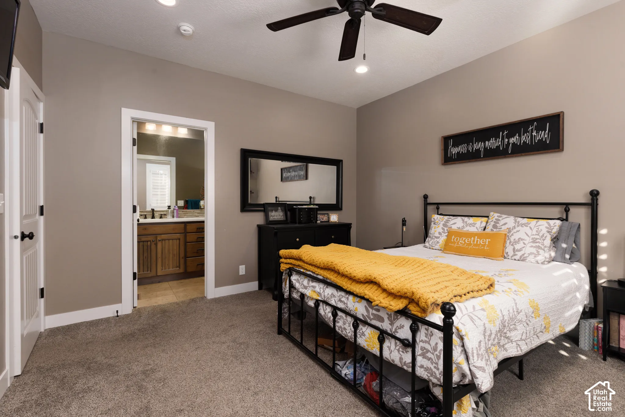 Neutral-toned bedroom featuring carpeted flooring, a ceiling fan with light fixture, and a doorway to a bathroom with wood-finish cabinetry