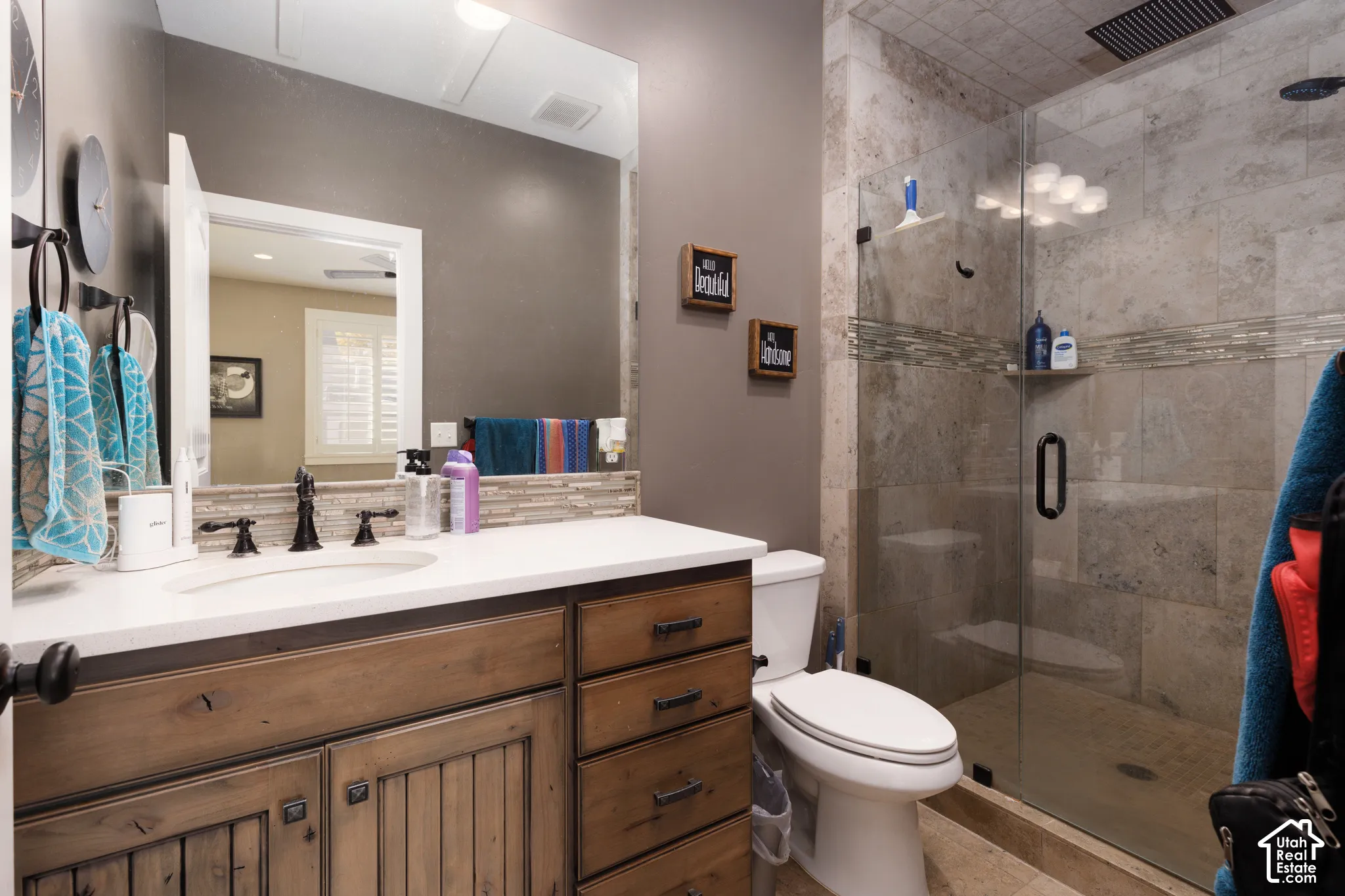 Bathroom featuring a wood-finish vanity with a light-toned countertop, large wall-mounted mirror, glass-enclosed shower with stone-style wall tile, and a decorative tile backsplash
