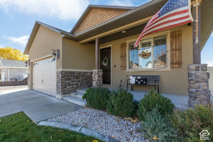 Exterior facade featuring tan stucco siding, stone veneer accents, shingle-style gable detailing, a covered front porch with timber posts, and decorative wooden shutters