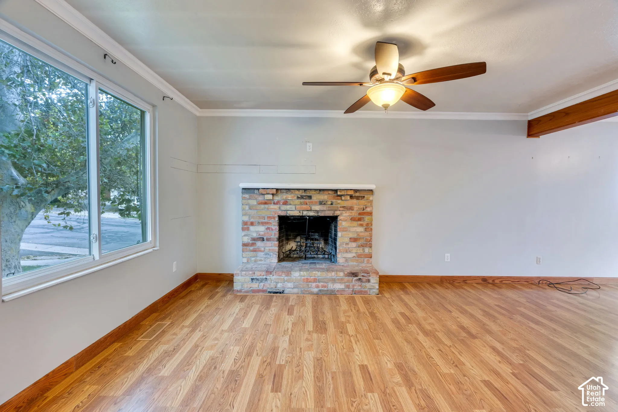 Brick fireplace featuring a hearth and mantle, wood-finish flooring, ceiling fan with integrated lighting, crown molding, and large window