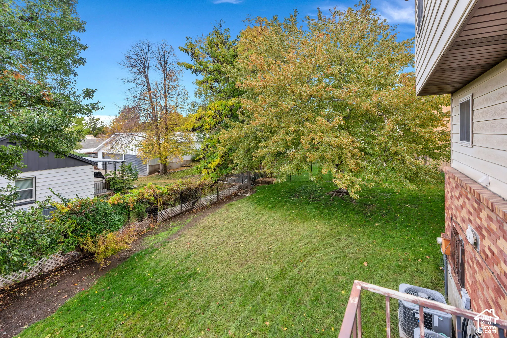 Exterior backyard area featuring a grass lawn, mature trees, lattice fencing, and a partial brick and siding fa