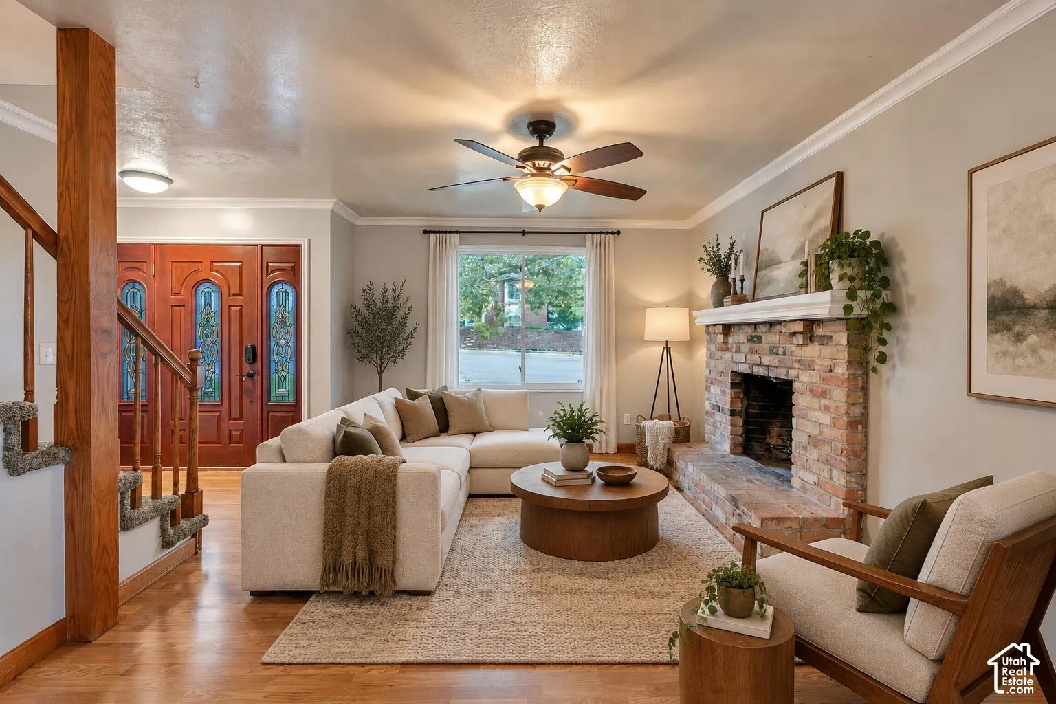 Living area featuring a masonry fireplace with a raised brick hearth and wood mantel, wood-finish flooring, crown molding, a central ceiling fan, and a wood-paneled entry door with decorative glass inserts
