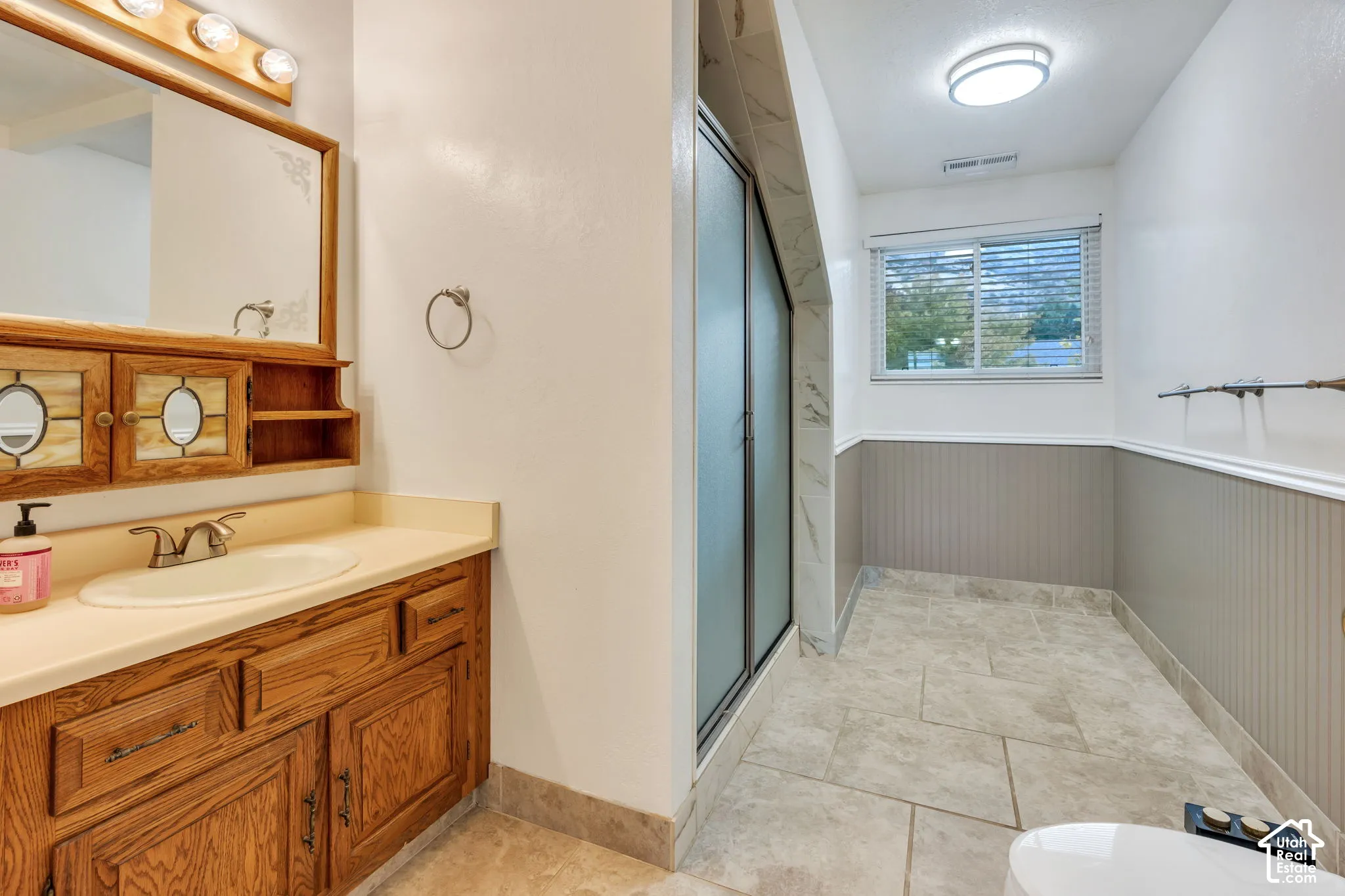 Bathroom featuring wood cabinetry with built-in shelving, tile flooring, glass-enclosed shower, wainscoting, and a sliding window