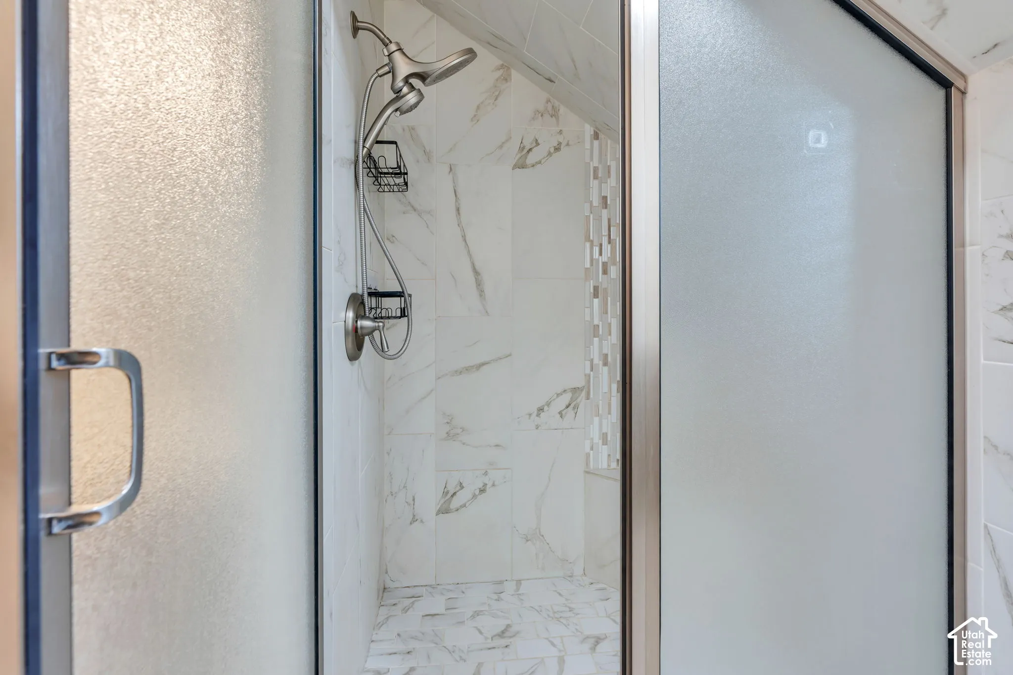Shower featuring full-height large-format tile with neutral veining, a brushed metal adjustable handheld showerhead, a built-in wire caddy, and a frosted glass sliding door with a brushed metal handle