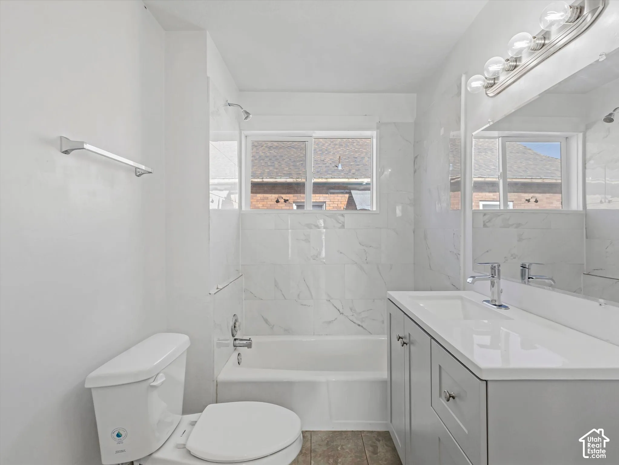 Bathroom featuring stone-finish wall tile, white soaking tub, light gray cabinetry with integrated white countertop, wall-mounted vanity mirror, and polished chrome fixtures