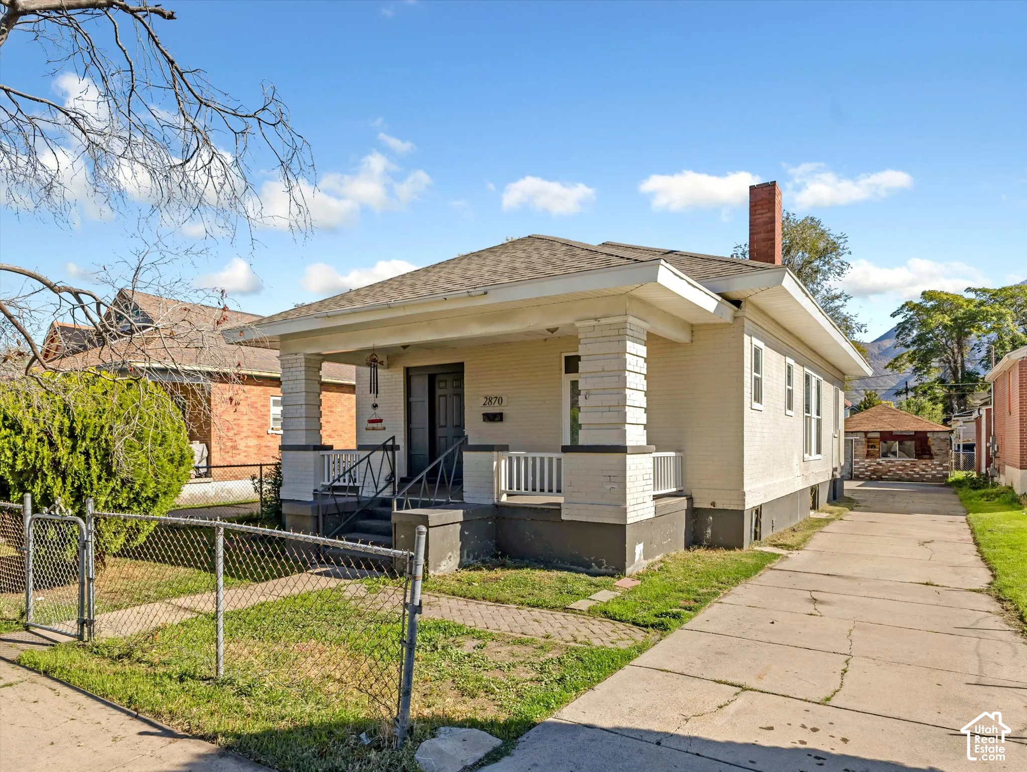 Bungalow exterior featuring light-colored brick siding, a covered front porch with tapered columns, a concrete driveway, and an exposed brick chimney