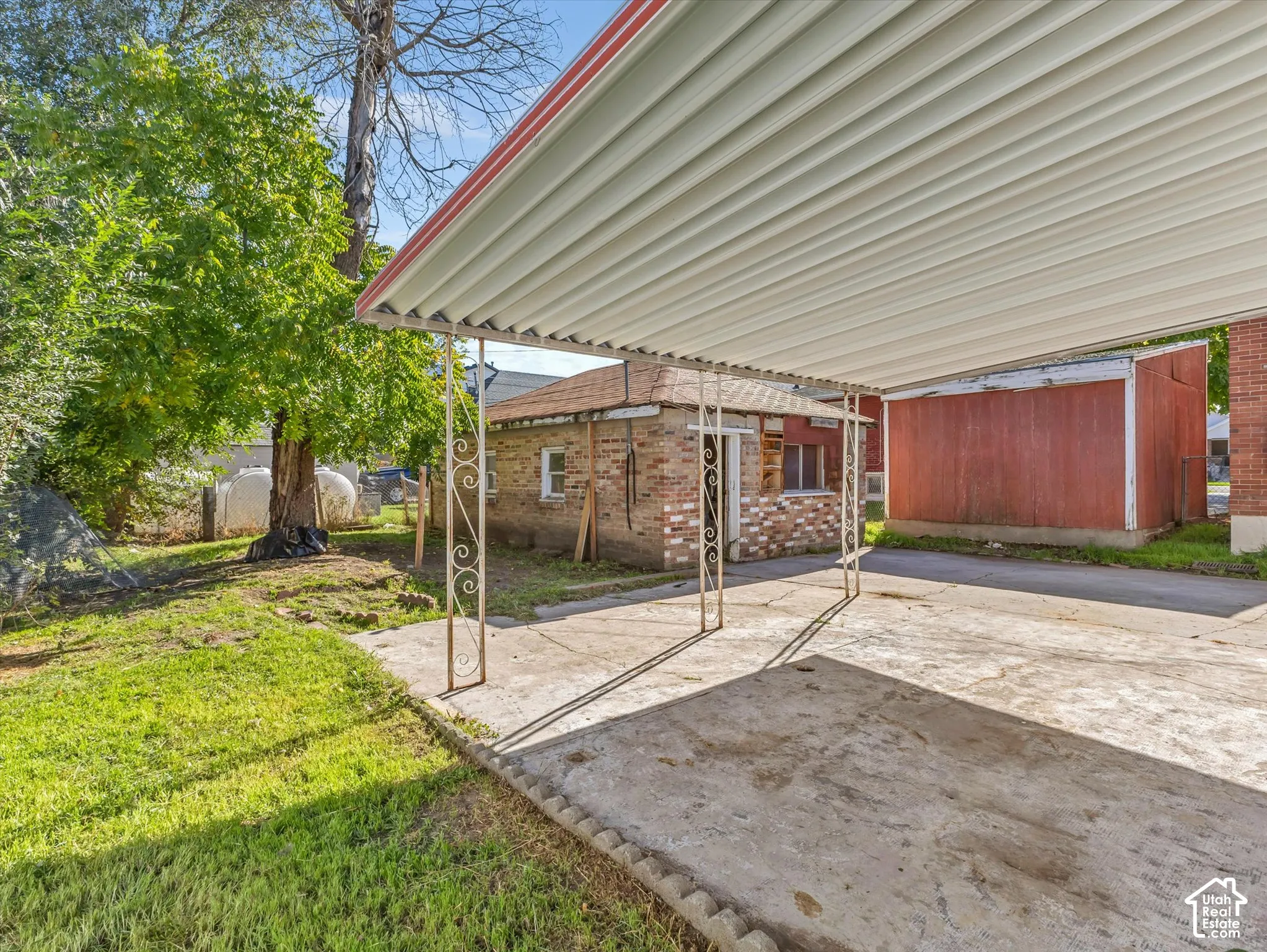Covered carport with ornamental metal support pillars and a concrete pad, accompanying a brick outbuilding and a detached storage structure on a grass-covered lot